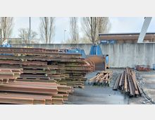 Ein Industriehof mit gestapelten Metallschienen, einer blauen Industriemaschine im Hintergrund und einer Betonwand. Bäume und ein Gebäude sind hinter der Wand sichtbar.