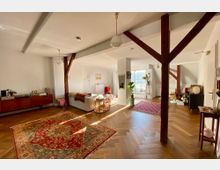 Offener Wohnraum mit weiß gestrichenen Wänden, Holzbalken und Parkettboden im Fischgrätmuster. Der Raum ist mit Teppichen, einem Sofa, Pflanzen und einer Schiebetür zu einem Balkon eingerichtet, wodurch viel Licht einfällt.