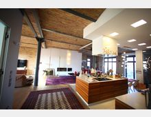 Offener Wohnraum in einem Loft mit Ziegelwänden, sichtbaren Metallträgern und Holzboden. Der Raum zeigt eine moderne Küche mit Holzfronten und Kochinsel, einen Wohnbereich mit lila Teppich, einer Schaukel und einem Kamin sowie große Fenster, die viel Tageslicht einlassen.