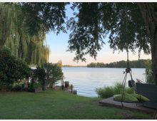 Gartenbereich mit Blick auf einen See, umgeben von Bäumen und Sträuchern. Im Vordergrund steht ein hängender Korbstuhl auf einer Holzbühne, neben Rasenfläche und einem Weidenbaum, der über das Wasser ragt.