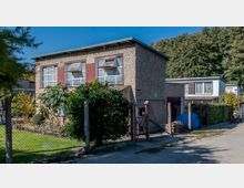 Zweistöckiges Wohnhaus mit grober Putzfassade, großen rechteckigen Fenstern und flachem Dach, umgeben von einem Zaun. Im Vorgarten befinden sich Sträucher und Blumen, im Hintergrund weitere ähnliche Gebäude sowie Bäume.