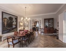 Wohnzimmer mit klassischer Einrichtung, darunter ein großer Holzesstisch mit Stühlen, ein Kronleuchter, eine Vitrine und eine Bank unter einem großen Gemälde. Hölzerner Fischgrätboden, helle Wände und Doppeltüren, die einen Blick in einen angrenzenden Raum mit großen Fenstern ermöglichen.