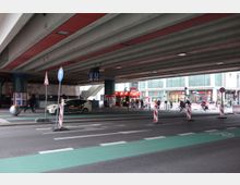Straßenansicht unter einer großen, betonierten Autobahnbrücke mit deutlichen roten Akzenten an der Decke. Im Vordergrund sind Straßenmarkierungen und Baustellenabsperrungen, während im Hintergrund ein U-Bahn-Eingang mit einem „U“-Schild sowie ein Imbissstand und umliegende Geschäfte sichtbar sind.