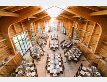 Holzinnenraum einer Veranstaltungshalle mit hohen Decken und sichtbarer Holzkonstruktion. Mehrere lange Holztische mit gedeckten Plätzen und dekorativen Elementen sind in Reihen aufgestellt; Tageslicht fällt durch große Fenster auf der linken Seite und ein Oberlicht ein.