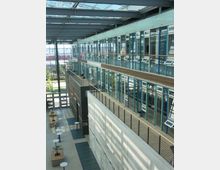 Offener Innenraum eines modernen Büro- oder Universitätsgebäudes mit mehreren verglasten Etagen. Große Fensterfronten, Metallgeländer und eine Glasdachkonstruktion lassen viel Tageslicht herein; unten befinden sich Sitzbänke und kleine Tische.