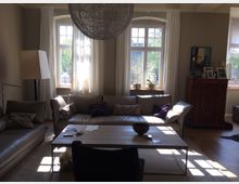 Wohnzimmer mit großer Fensterfront, zwei gepolsterten Sofas und einem langen Couchtisch aus Holz. Der Raum ist in neutralen Tönen gehalten, mit weißen Vorhängen, einer Stehlampe und dekorativen Gegenständen auf einem hohen Holzschrank.