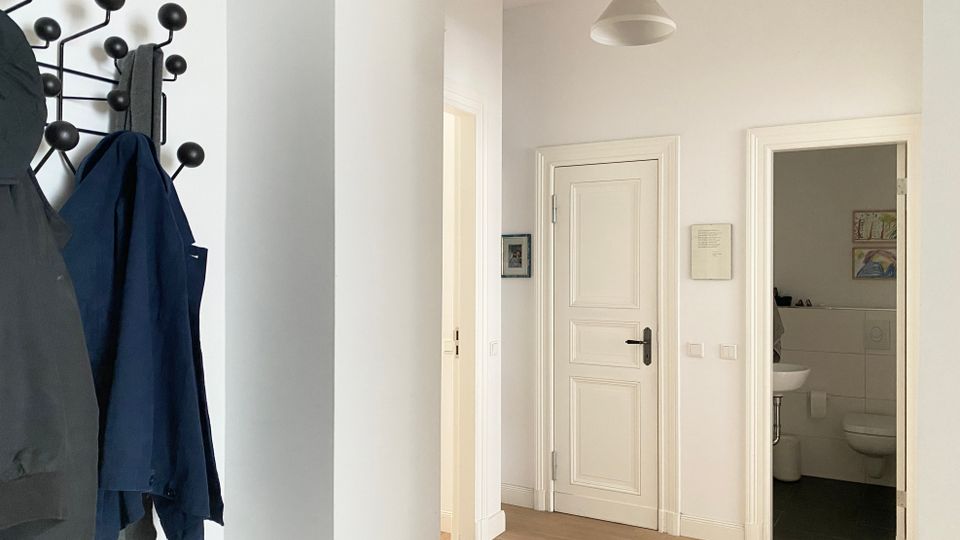 Heller Wohnungsflur mit weiß gestrichenen Wänden, Holzboden und einer Deckenleuchte, mit mehreren weißen Zimmertüren. Links hängt eine Garderobe an einer schwarzen Wandhakenleiste, rechts ist eine geöffnete Tür zu einem kleinen Bad mit Waschbecken und Toilette zu sehen.