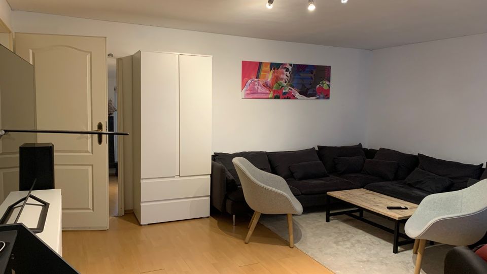 Wohnzimmer mit heller Holzoptik-Bodenfläche, großer schwarzer Eckcouch und Couchtisch auf einem hellen Teppich. An der weißen Wand hängt ein farbiges Bild, daneben stehen ein weißer Schrank und eine Tür; an der Decke sind Spot-Schienenleuchten montiert.