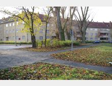 Wohnblock im Stil der 1920er- bis 1930er-Jahre mit gelblichen Fassaden, roten Ziegeldächern und zahlreichen Fenstern, umgeben von Laubbäumen. Im Vordergrund verlaufende Gehwege, ein kleiner parkähnlicher Bereich und verstreutes Herbstlaub.