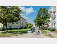 Wohnstraße in einem Wohngebiet mit mehrstöckigen, beige-weißen Gebäuden auf beiden Straßenseiten, bepflanzten Grünflächen, parkenden Autos und Bäumen; eine Person fährt mit dem Fahrrad, und Straßenschilder sind sichtbar.