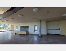 Ein leerer Innenraum mit gefliestem Boden, abgehängter Decke mit runden Leuchten und teilweise abblätternder Farbe. Im Vordergrund ein Empfangstresen, dahinter eine Garderobe; große Glasfenster lassen Tageslicht herein.