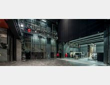 Bühnenhintergrund eines Theaters mit sichtbaren technischen Einrichtungen, wie Bühnenbeleuchtung, Hebebühnen und seitlich gelagertem Equipment. Die Szenerie ist durch schwarze Vorhänge, hohe Wände und Metallgerüste geprägt.