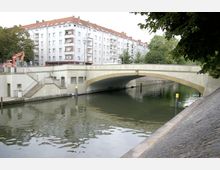 Brücke über einen Kanal in einem städtischen Umfeld mit Wohngebäuden im Hintergrund. Der Kanal ist von befestigten Ufern und Bäumen gesäumt, und eine Treppe führt von der Brücke hinunter zum Wasser.