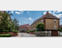 Wohnstraße mit zweigeschossigen Reihenhäusern, die braune Putzfassaden und rote Ziegeldächer haben. Die Straße ist von kleinen Vorgärten, Bäumen und Hecken gesäumt, bei sonnigem Himmel mit wenigen Wolken.