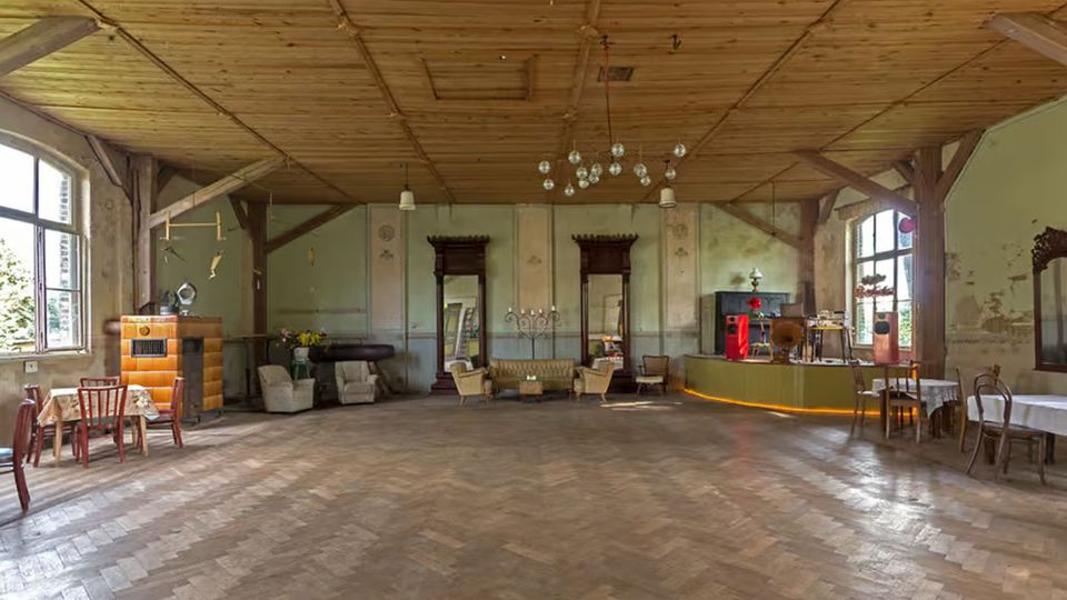 Großer, leerer Saal mit Fischgrät-Parkettboden, holzvertäfelter Decke und sichtbaren Holzbalken sowie hohen Fenstern an den Seiten. Entlang der Wände stehen vereinzelte Vintage-Möbel und Dekoration, darunter Stühle, Sessel und ein kleiner Bartresen.