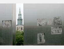 Blick durch eine Metallplatte mit eingravierten Schwarz-Weiß-Bildern von historischen Szenen, im Hintergrund der Turm einer Kirche mit grünlicher Spitze und einer Uhr. Vordergrund und Hintergrund sind durch die Öffnung der Metallstruktur optisch verbunden.