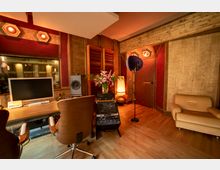 Aufnahmestudio mit Holz- und roten Akustikpaneelen, einem Schreibtisch mit Monitor und Lautsprecher, professionellem Tonmischpult, Mikrofonständer, einer Sitzgarnitur und einer Vase mit Blumen. Die Beleuchtung ist warm, und der Boden besteht aus Holz.