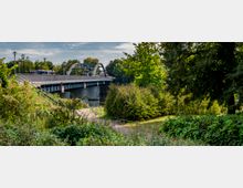 Eine Brücke mit einem breiten Bogen führt über einen Fluss, daneben Busse und Fußgänger. Im Vordergrund befinden sich grüne Büsche, Bäume und ein Weg in einer parkähnlichen Landschaft.