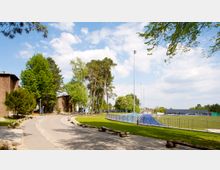 Fußweg in einer parkähnlichen Umgebung mit Bäumen und Grünflächen; links ein Backsteingebäude, rechts ein umzäuntes Sportfeld mit Kunstrasen. Im Hintergrund sind moderne, flache Gebäude und ein blauer Himmel mit Wolken zu sehen.