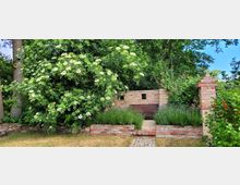 Ein kleiner Gartenplatz mit einer Holzbank, umgeben von einer niedrigen Steinmauer aus rotem Backstein. Lavendel und üppige grüne Sträucher, darunter ein blühender Holunderbaum, umrahmen den Bereich, mit einem Weg aus Pflastersteinen, der zur Bank führt.