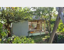 Kleines Gartenhaus mit Holzrahmen und großen Fenstern, umgeben von dichtem Laub und Obstbäumen. Im Inneren sind Regale, Utensilien und Sitzgelegenheiten sichtbar; die Umgebung wirkt natürlich und dicht bepflanzt.