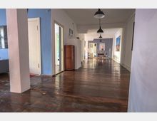 Langer, heller Flur mit Holzboden und weißen Wänden, teilweise in Blau gestrichen. Im Hintergrund stehen ein Tisch mit Stühlen, Wandleuchten und gerahmte Bilder an der Wand, während Türen in angrenzende Räume führen.