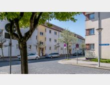 Wohnstraße mit Mehrfamilienhäusern in hellen Farben, teils verputzt, teils mit Klinkerelementen. Im Vordergrund stehen Bäume, Straßenschilder, parkende Autos und ein Straßenname („Havelberger Straße“) sind sichtbar.