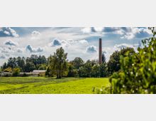 Weite Wiese mit grünem Gras und vereinzelten Bäumen im Vordergrund, dahinter eine Baumreihe, ein Gebäude mit Flachdach und ein hoher Ziegelschornstein. Am Himmel sind Wolkenformationen zu sehen.