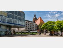 Straßenszene in einer Stadt mit moderner Glasfassade eines Bürogebäudes links, einer historischen Kirche mit rotem Dach und Turm im Hintergrund, sowie einer grünen Straßenbahn auf den Schienen im Vordergrund. Der Bereich ist von Bäumen, Fußgängern und Fahrradfahrern belebt.