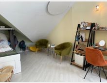 Zimmer mit schräger Dachdecke und Holzboden, ausgestattet mit einem Bett auf der linken Seite, einer grünen Samtsitzgruppe, einem gelben Sitzmöbel, einem kleinen Tisch, einem Regal mit Büchern und Dekorationen sowie einem Schreibtisch mit orangener Stuhl. Die Wände sind hellgelb gestrichen, und eine Wandlampe sorgt für Beleuchtung.