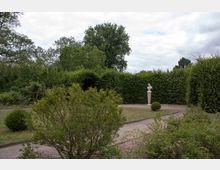 Ein gepflegter Garten mit einer sandigen Gehwegfläche führt zu einer weißen Büste auf einem Sockel, umgeben von dichten Hecken und Bäumen im Hintergrund. Im Vordergrund befinden sich Sträucher und kleine Büsche.