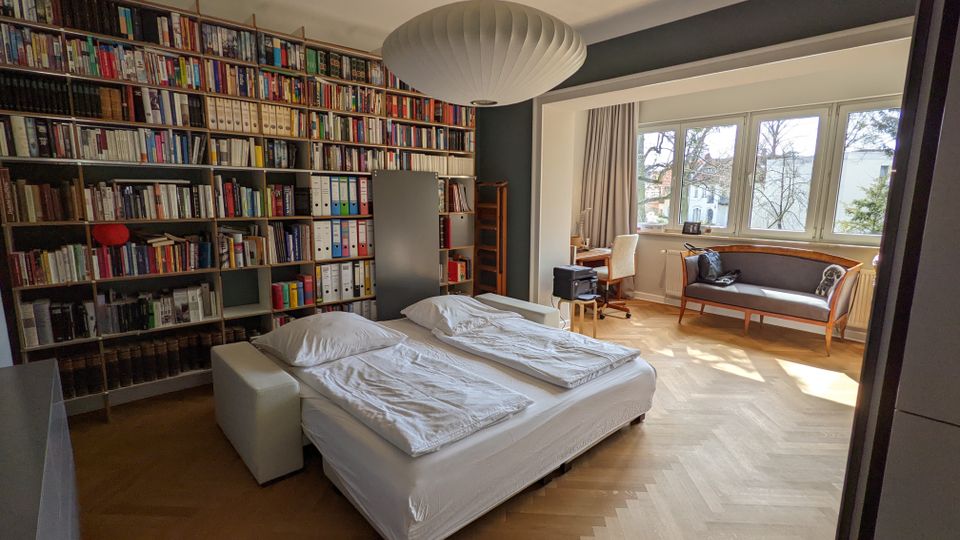 Helles Schlafzimmer mit Parkettboden, Doppelbett und einer wandfüllenden Bücherregalwand. Große Fensterfront mit Sitzbank/kleinem Sofa und Schreibtischbereich sowie einer großen Deckenleuchte.