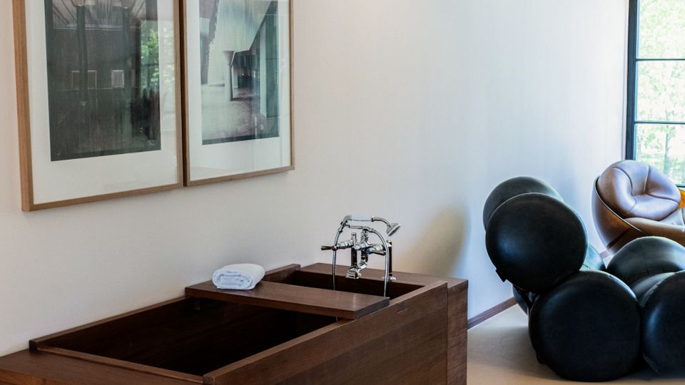 Modernes Badezimmer mit freistehender Holzbadewanne, zwei gerahmten Bildern an einer schlichten weißen Wand und sichtbaren weißen Deckenbalken. Im Hintergrund befinden sich kugelförmige Möbelstücke und ein Fenster mit schwarzem Rahmen.