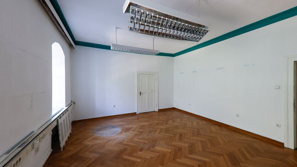 Leerer, heller Innenraum mit Fischgrät-Parkettboden, weißen Wänden mit türkisfarbener Zierleiste und großen rechteckigen Deckenleuchten. Links befindet sich ein hohes Fenster über einem Heizkörper, gegenüber eine weiße Tür und rechts ein weiterer Türdurchgang.