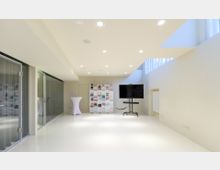 Heller, moderner Veranstaltungsraum mit glänzendem, weißem Boden, einer Glaswand links, einem weißen Regal mit Büchern an der Rückwand sowie einem großen Flachbildschirm auf einem mobilen Ständer rechts. Oben sorgen eingelassene Deckenleuchten für gleichmäßige Beleuchtung.