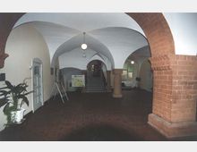 Gewölbter Innenraum eines historischen Gebäudes mit roten Sandsteinsäulen und Spitzbögen. Im Hintergrund befinden sich eine Treppe und ein Infostand, links und rechts Türen und Pflanzen.