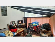 Überdachter Raum mit Glasdach und beiger Stoffabtrennung, gefüllt mit verschiedenen Gegenständen wie Fahrrädern, einem Schlauchboot, einem Holztisch mit Haushaltsartikeln, einem alten Holzschrank und gestapelten Büchern. Links sind Glasbausteine in der weißen Wand eingebaut.