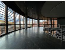 Großer, moderner Innenraum mit schwarzen Böden und einer gebogenen Glasfassade, die durch horizontale Holzlamellen beschattet wird. Eine Metallgeländerbrüstung verläuft entlang eines offenen Bereichs, und durch die Fenster ist eine urbane Außenlandschaft sichtbar.