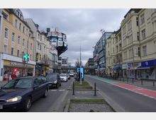 Straßenszene mit mehrstöckigen Gebäuden auf beiden Seiten, darunter Geschäfte wie eine Apotheke und eine C&A-Filiale. Eine Schwebebahn fährt oberhalb der Straße, die durch eine Fahrradspur und parkende Fahrzeuge aufgeteilt ist; Passanten und Autos beleben den Bereich.