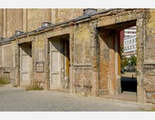 Ziegelsteinfassade einer Ruine mit offenen Torbögen und verblassten Putzresten, umgeben von einem gepflasterten Platz. Im Hintergrund sind moderne Gebäude und ein "S" Symbol für eine S-Bahn-Station zu sehen.