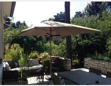 Terrasse im Grünen mit Sitzmöbeln, großem Sonnenschirm und Pflanzen. Im Vordergrund steht ein Holztisch, im Hintergrund sind dicht bepflanzte Beete und eine Steinmauer sichtbar.