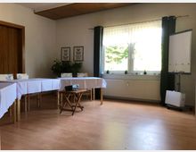 Ein heller Seminarraum mit Holzboden, Tischen mit weißen Tischdecken, einem kleinen Holztisch mit Beamer in der Mitte, einem Flipchart und einem großen Fenster mit dunklen Vorhängen. Pflanzen und Schmetterlingsbilder schmücken den Raum dezent.