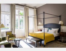 Helles Schlafzimmer mit einem Himmelbett und gelber Decke, flankiert von klassischen Nachttischen mit Lampen. Der Raum hat hohe Fenster mit weißen Vorhängen, durch die Tageslicht einfällt, sowie eine Sitzgruppe mit grünem Sofa, gemustertem Sessel und kleinem Glastisch.