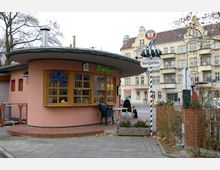 Rundliches Gebäude mit einer rosafarbenen Fassade und einem großen Fenster mit Holzrahmung, das als Verkaufstresen dient. Ein Schild mit der Aufschrift "Biergarten" steht daneben, umgeben von Pflanzenkübeln und mit mehrstöckigen Altbauten im Hintergrund.