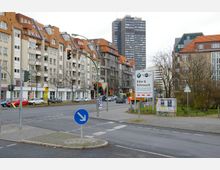 Straßenkreuzung in einer Stadt mit mehrstöckigen Wohn- und Geschäftsgebäuden im Hintergrund, teilweise mit roten Ziegeldächern. Im Vordergrund sind Verkehrszeichen, Ampeln, und Werbetafeln zu sehen, umgeben von parkenden Autos und kahlen Bäumen.