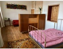 Schlicht eingerichtetes Schlafzimmer mit Holzboden und Teppich, bestehend aus einem Metallbett mit rot-weiß karierter Bettwäsche, einem Holzschrank, einer roten Couch, einem kleinen Beistelltisch mit Lampe, sowie einer abstrakten Wanddekoration. Eine eingefasste, offene Holzluke befindet sich in der Wand.