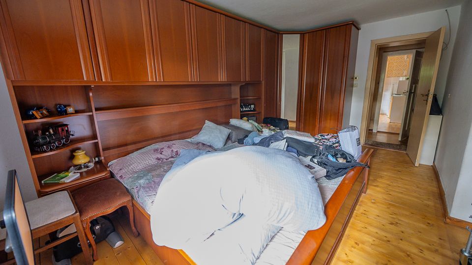 Schlafzimmer mit großem Holzbett und wandfüllendem Holzschrank, der ein integriertes Regal und eine Ablage am Kopfteil bildet. Holzdielenboden und offene Tür zu einem Flur mit Blick in einen weiteren Raum.