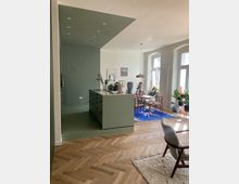 Moderne Wohnküche mit grünem Einbauschrank und Arbeitsinsel, die in einen offenen Essbereich mit Holztisch, Stühlen und blauem Teppich übergeht. Der Raum ist hell, mit großen Bogenfenstern, Pflanzen und Holzboden gestaltet.