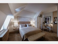 Ein elegantes Schlafzimmer mit einem Bett in der Mitte, flankiert von zwei Nachttischen mit Lampen. Der Raum hat schräge Decken, ein kunstvoller Wandrelief hinter dem Bett, helle Holzmöbel, einen Schreibtisch mit Stuhl und eine warme Beleuchtung.