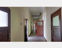 Ein schmaler Flur mit cremefarbenen Wänden und dunklen Holztüren mit Glasfenstern, die teils geöffnet sind. Am Ende des Flurs befindet sich eine Ausschilderung und ein Hinweis auf einen Ausgang.